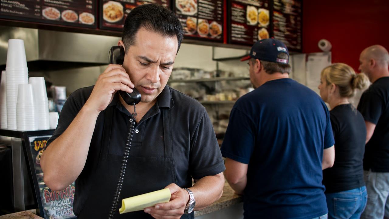 Empleado de restaurante contestando el teléfono con una libreta mientras clientes esperan en el mostrador